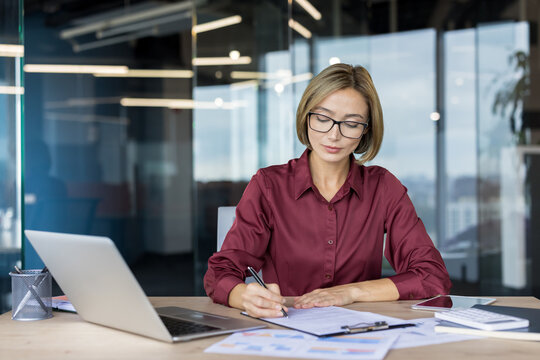 Young businesswoman reviewing financial documents and writing notes at desk with laptop, demonstrating concentration and dedication in a professional corporate environment