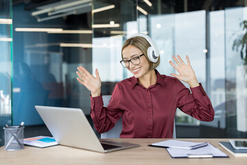 Young woman in office wearing headphones, smiling and dancing at her desk while listening to music,...