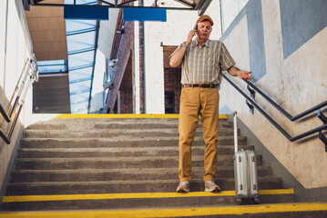 Fototapeta premium Senior man is standing at railway station and talking on mobile phone. He is distraught. Senior people traveling.