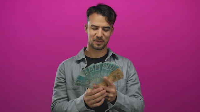 Young hispanic man holds brazilian reais cash over isolated pink background, showcasing wealth and confidence.