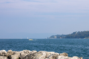 Lake seascape with rocky shoreline in foreground. Motorboat and sailboat on water, distant coastline on right. Wide horizontal composition with copy space sky