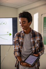 African American male holding tablet with diagrams in class, monitor showing graph and whiteboard