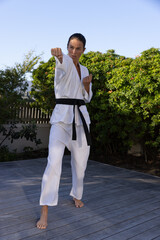 Karate gi delivering punch on wood-plank deck bordered by shrubs under clear sky, copy space