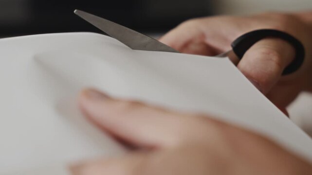 Close up shot of male hands cutting white paper with scissors on a table. Camera focuses on fingers and blade movement, shallow depth of field, calm home atmosphere. High quality 4k footage