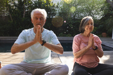 Senior couple meditating cross-legged on yoga mats on wooden deck beside pool wearing sport watches