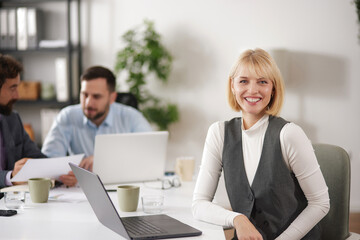 Obraz premium A business team collaborates on ideas at a table in a bright office. There are laptops, documents, and cups on the table. One member smiles while others discuss.