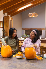 Naklejka premium Diverse mother and teen daughter scooping pumpkin pulp and seeds, carving pumpkins in dining room