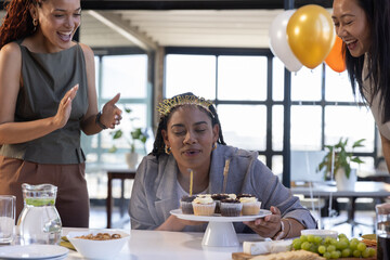 Diverse coworkers celebrating birthday by cake stand with cupcakes and balloons at open plan office