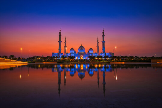 A beautiful reflection of a Grand Mosque is seen in the water. The sky is a deep purple and the water is calm