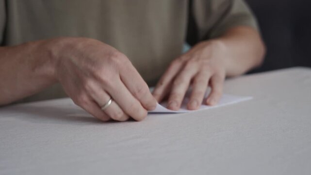 Medium shot slightly above table height showing an adult man forming a narrow edge of a paper airplane. Camera captures finger precision and careful folding motion. High quality 4k footage
