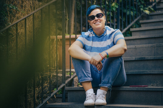 Smiling non-binary person with blue hair and sunglasses sitting on outdoor stairs, representing gender identity and queer visibility in daily life