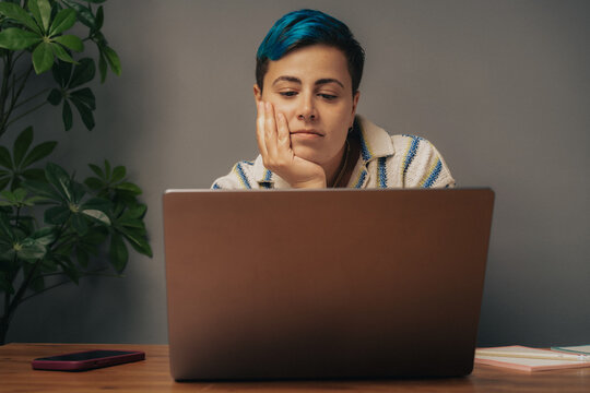 Frustrated non-binary student with blue hair looking at laptop screen during online education class at home