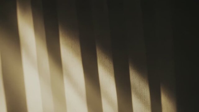 Warm golden light entering through a curtain or window forms abstract parallel striped shadows and gradients on a textured wall, creating a serene and tranquil background