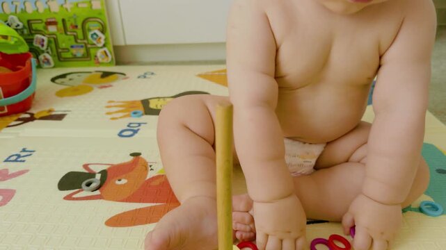 Baby in diaper sits on colorful play mat, stacking rings on a wooden post, showcasing hand-eye coordination and playful exploration of shapes and colors