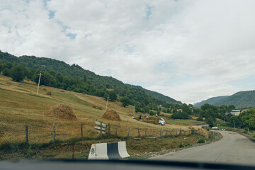 Obraz premium Winding rural road through rolling hills and open fields, fences along the roadside, cloudy sky, a peaceful countryside landscape viewed from a car