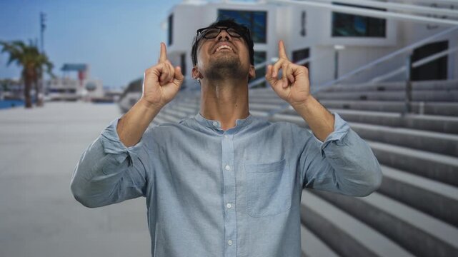 Young hispanic man outdoors pointing upwards with a joyful expression near modern architecture and palm trees under a clear sky conveying excitement and positivity in a public setting.