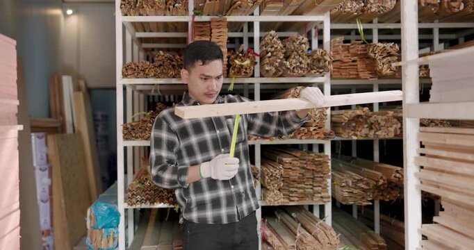 Asian adult man craftsman measures wooden board width while standing among wood planks stacked on shelves during artisan wood selection process for interior decoration raw materials
