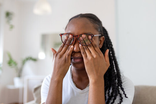 Tired Black woman taking off glasses to rub her eyes due to digital screen strain. Concept of work burnout, headache, fatigue, and vision problems while working from home.