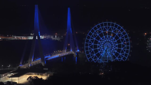 Integration Bridge with glowing blue lights and large illuminated Ferris wheel near the Paran&aacute; River in Foz do Igua&ccedil;u and Ciudad del Este Triple Frontera landmark. Aerial view at night.