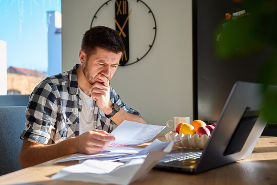 Worried man checking bills and financial papers while sitting at table with laptop. Stressed person planning home budget. Concept of personal finance review, expense control and household budgeting