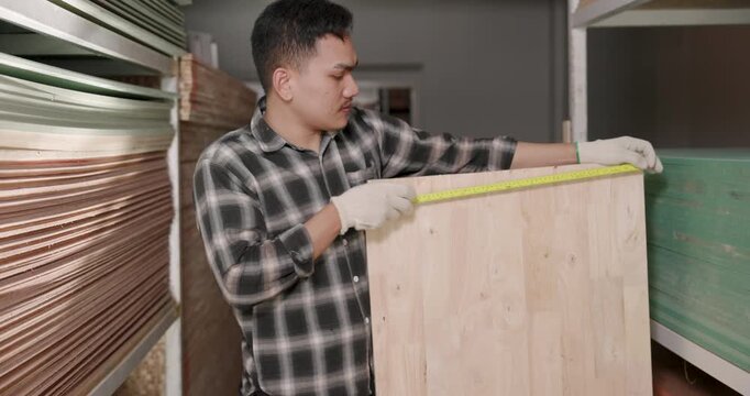 Asian adult male carpenter checks wooden board length using tape measure along shelf inside woodworking supply store, ensuring accurate sizing and material suitability for furniture making process