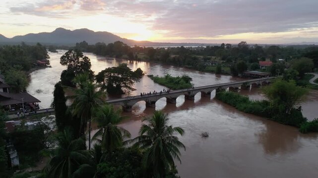 Aerial view of a stone bridge crossing a wide river at sunset in Don Det, Laos, surrounded by trees, palms, and village houses.