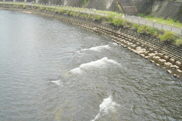 都市部を流れる穏やかな川の風景写真。護岸に並ぶ石とゆるやかな水の流れが、日常の中の自然や落ち着いた時間、散歩やリフレッシュをイメージさせる日本の街並みスナップ。