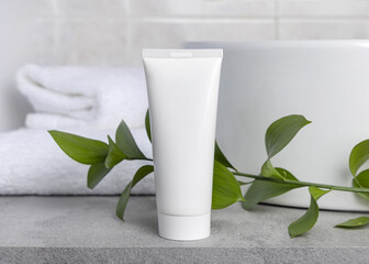 White blank cream tube near green leaves, basin and white bath towels on grey stone, mockup