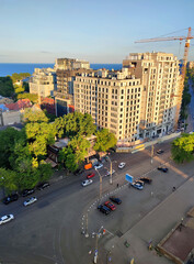 City street view from above. Large multi-story buildings, a square, green trees, an asphalt road for cars, moving cars and walking people on a sunny summer evening