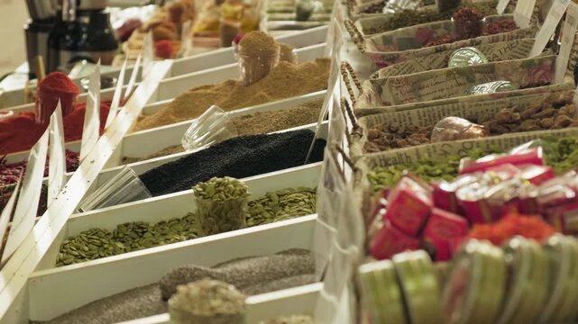 Hand selecting spices from market boxes, closeup of fingers picking lentils and herbs from white trays neat rows of pulses and colorful packs, vendor adjusting display, cinnamon sticks and jars