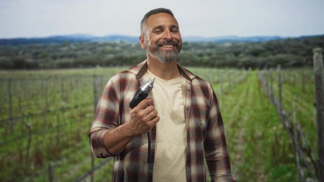 Man holds power drill to chest in vineyard rows, smiling with closed eyes and bare forearm visible while wearing flannel shirt and t shirt; pride.
