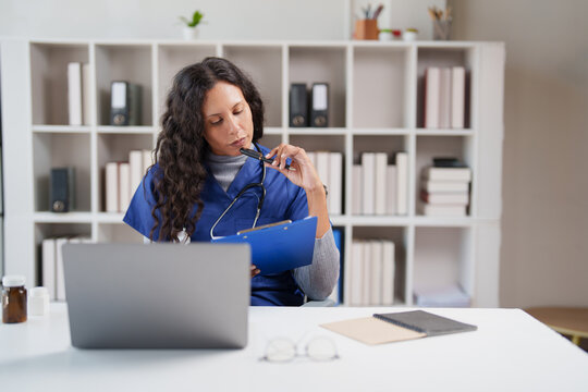 Female doctor or nurse in blue scrubs intently thinking and reviewing notes on clipboard, working at a desk with a laptop and stethoscope in a modern clinic office