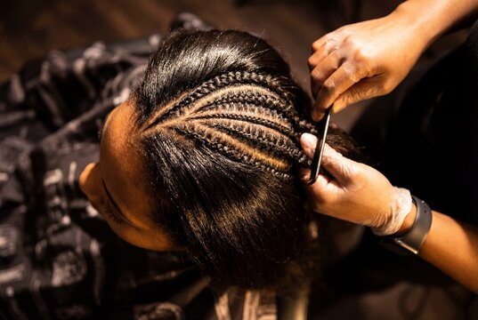 Hairdresser meticulously braids intricate cornrows on a woman dark hair with a comb