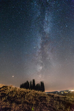 View of a striking celestial display of the Milky Way arches over a dark silhouette of trees on a hill under the night sky, Val d'Orcia, Tuscany, Italy.