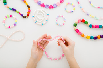 Girl holding pink necklace on light gray table background. Creating different beaded jewellery. Little child handcraft. Closeup. Point of view shot. Flat lay. Top down view.