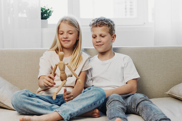 Obraz premium Two children sitting on couch playing with wooden mannequin, smiling and enjoying creative activity in bright living room with natural light and casual clothes.