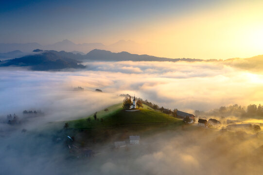 Aerial view of the church on the hill rises above the golden sea of mist, creating a serene and ethereal landscape, Skofja Loka, Slovenia.