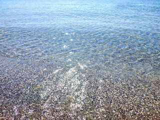 Clean transparent water on shallow sandy seashore with shells with shells on sunny summer day. Sun glare on surface of water. Small waves on sea coast. Nature backdrop. Natural background