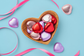 Chocolate hearts with colorful foil wrapped in heart wooden bowl with pink ribbon on blue background, Valentine's day celebration