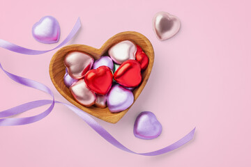 Heart chocolate with colorful foil wrapped in heart wooden bowl with purple ribbon on pink background, Valentine's day and love celebration, top view
