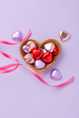 Valentine's day and love concept, chocolate hearts with colorful foil wrapped in heart wood bowl with pink ribbon on purple background, top view