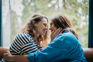 Fototapeta premium Lesbian couple sitting face to face on sofa touching each other’s faces, intimate moment at home