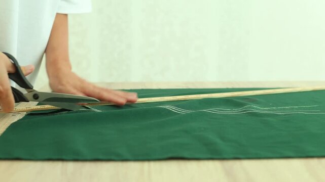 A person meticulously cuts dark green fabric with scissors, following a chalk line and using a ruler for precise, straight edges in garment making.