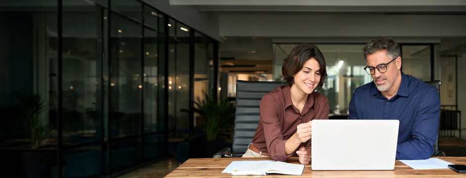 Female executive manager explaining corporate project plan to male colleague sitting at office desk. Business team of two busy professionals working together discussing ai solutions using laptop.