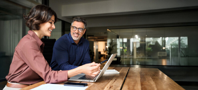 Business team two happy busy executives working together using laptop in office. Young business woman manager showing presentation to mature man investor, consulting client partner at work meeting.