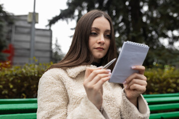 Adult girl writes notes in a notebook on a park bench