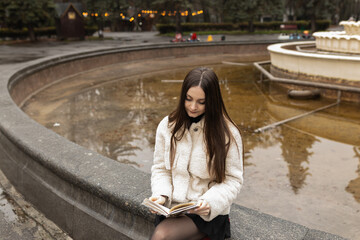 Adult girl is reading a book in a park near a fountain