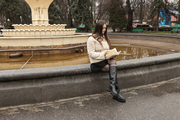 Adult girl is reading a book in a park near a fountain