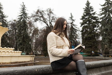 Adult girl is reading a book in a park near a fountain