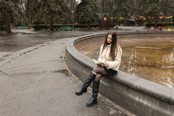 Adult girl is reading a book in a park near a fountain